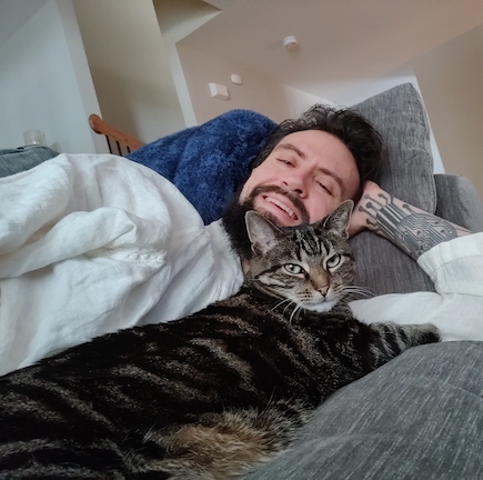 Alex is lounging behind his cat, wearing a white shirt. Both Alex and cat are looking at the camera, Alex with a big smile.