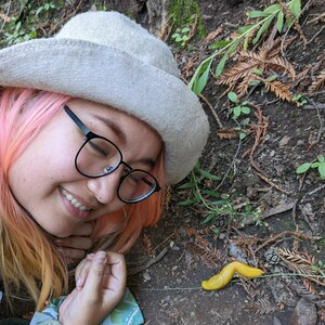 Jenni, a person with peach/pink colored hair, black glasses, and a hat is smiling next to a yellow slug.